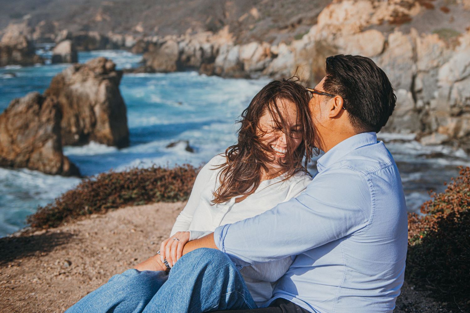 Couple sitting together on rocky coastal cliffs, smiling and embracing with ocean waves and sea stacks in the background.
