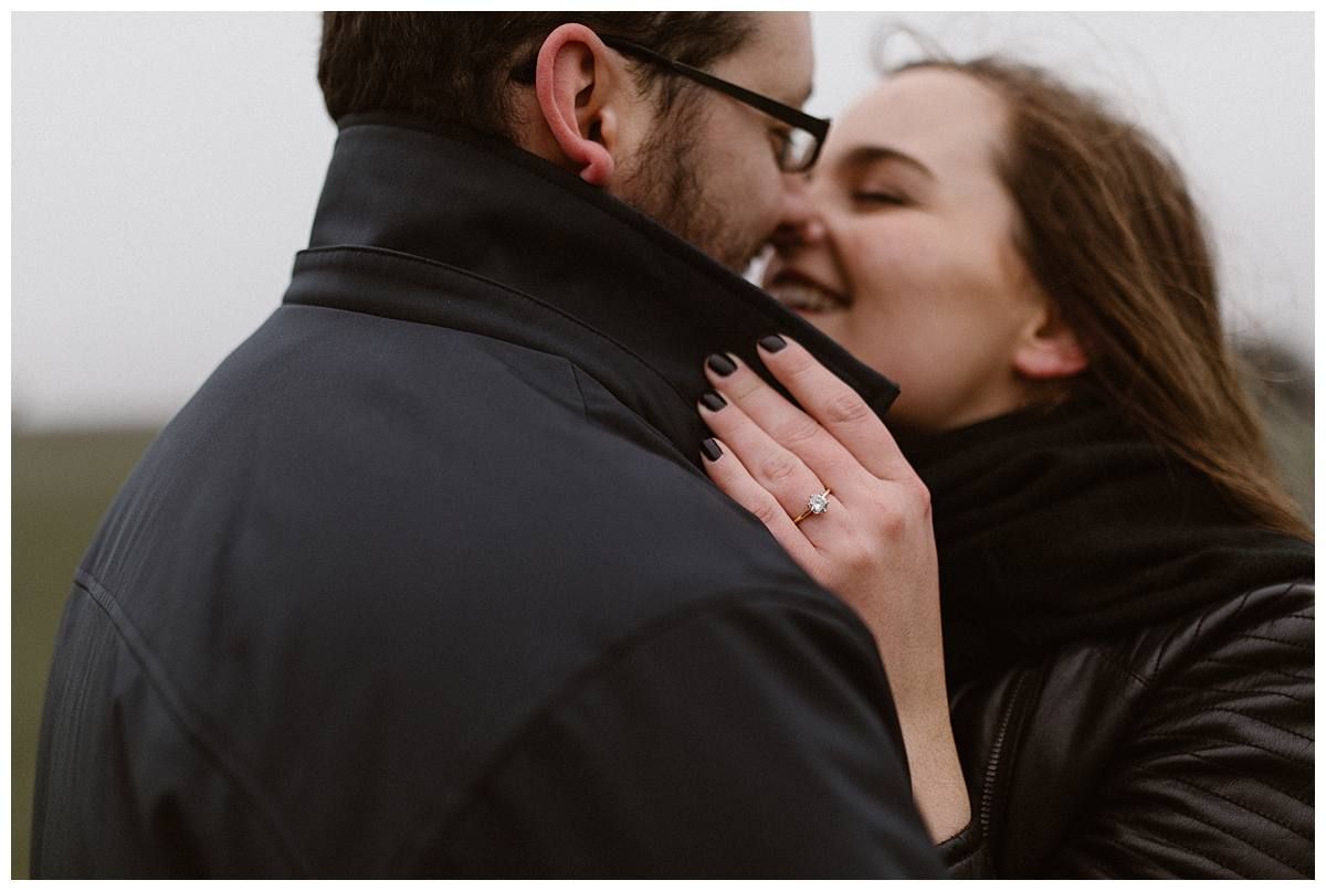 Couple sharing a joyful moment, woman smiling and showing engagement ring, wearing black leather jackets outdoors.