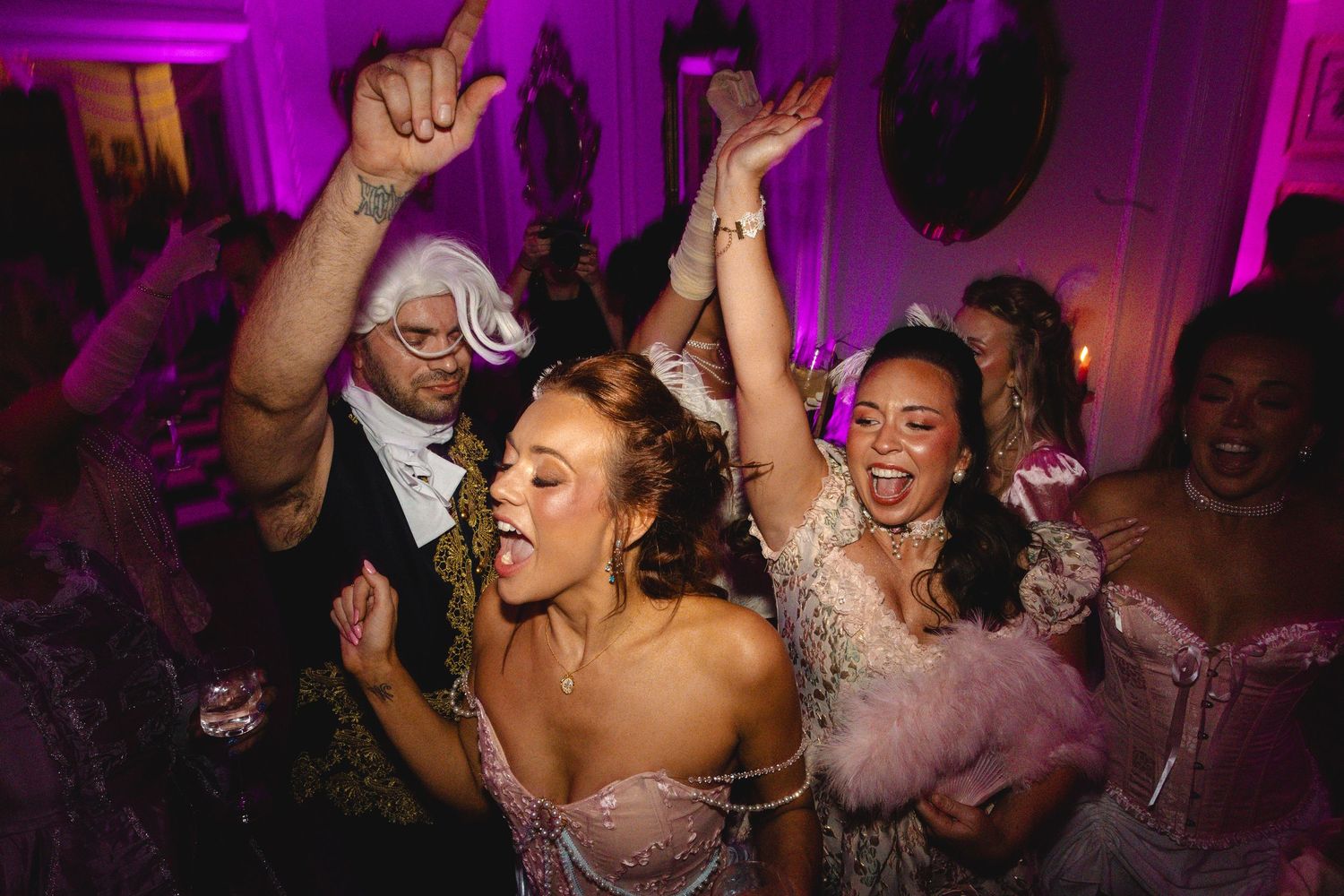 Guests in Marie Antoinette costumes dancing joyfully at a pink-lit masquerade ball.