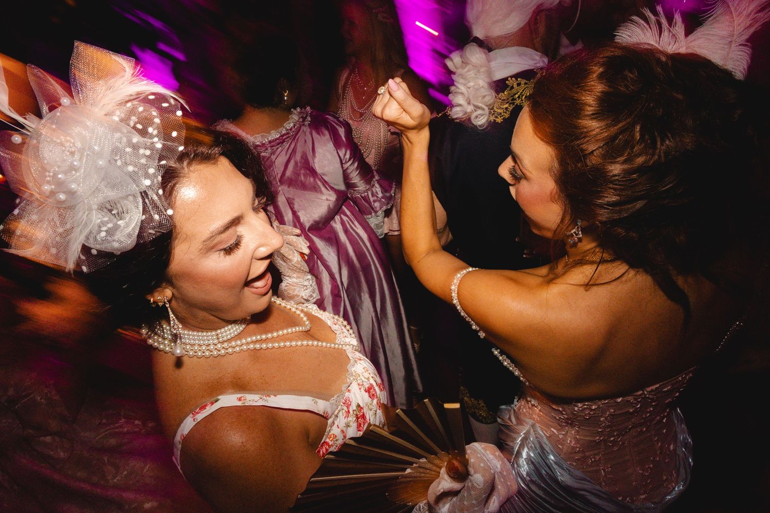 Two women in elegant attire dancing joyfully at a festive celebration with pink lighting.