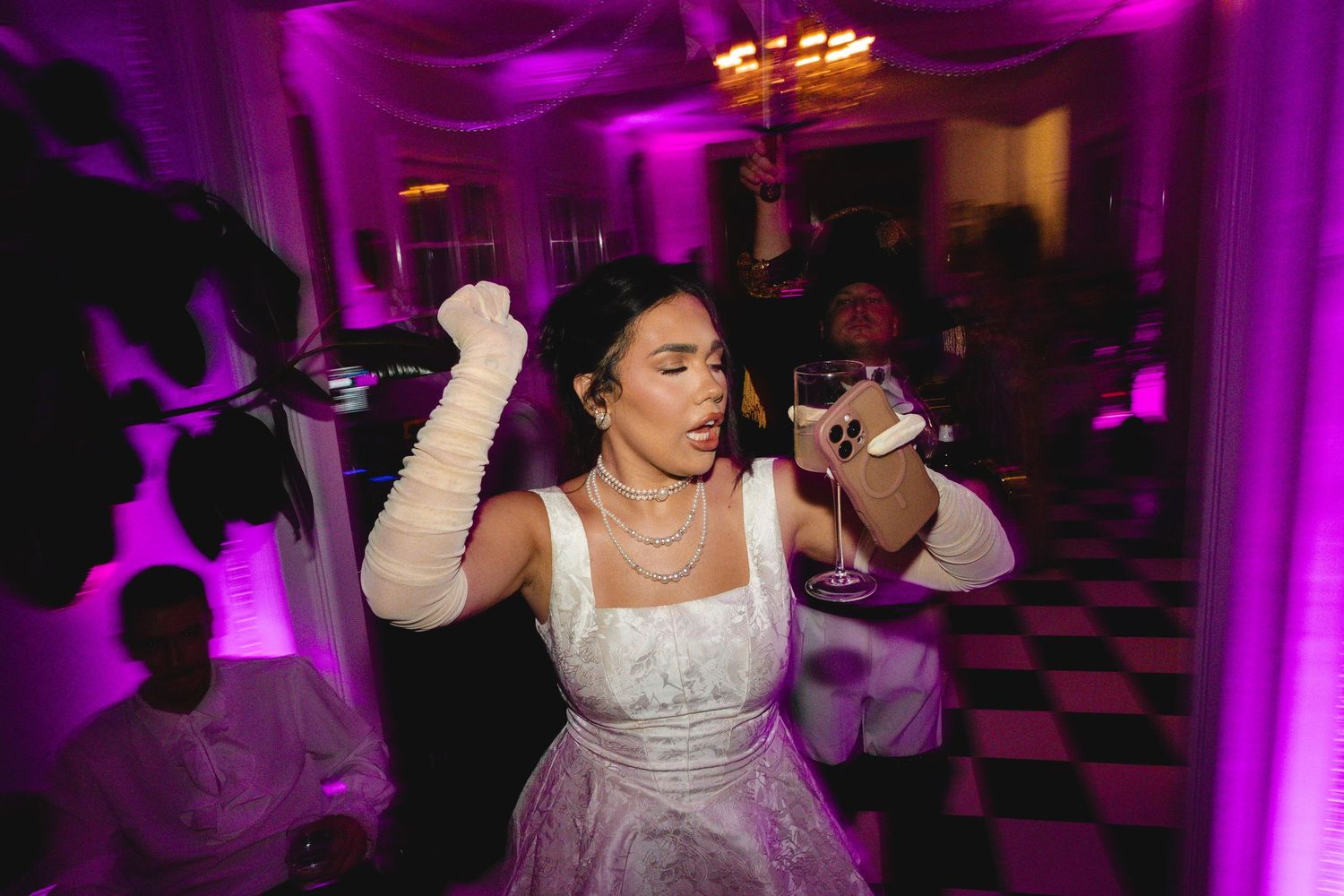 A bride in a white gown with long gloves dances energetically at a wedding reception with purple lighting.