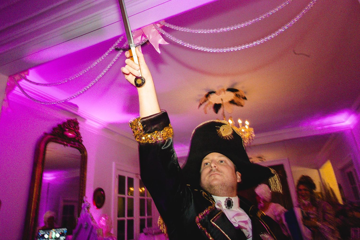 Man in Napoleon costume raises a sword at a glamorous pink-lit masquerade party.
