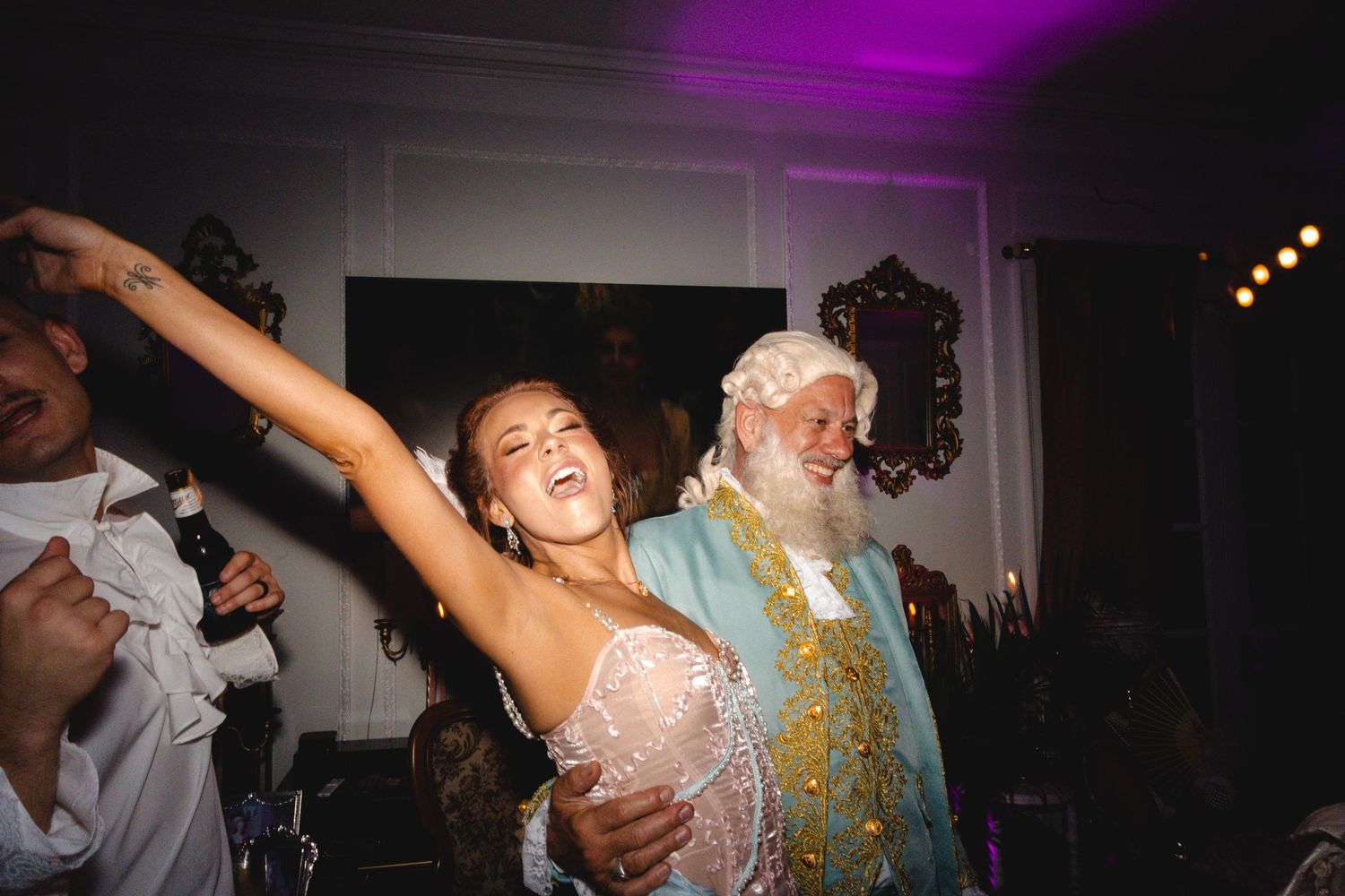 Bride and elderly man with white beard dancing joyfully at a wedding reception indoors.