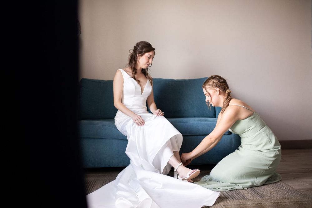 A bridesmaid in a mint green dress helps adjust a white wedding gown before the ceremony on a blue couch.  Iowa City wedding photographer.
