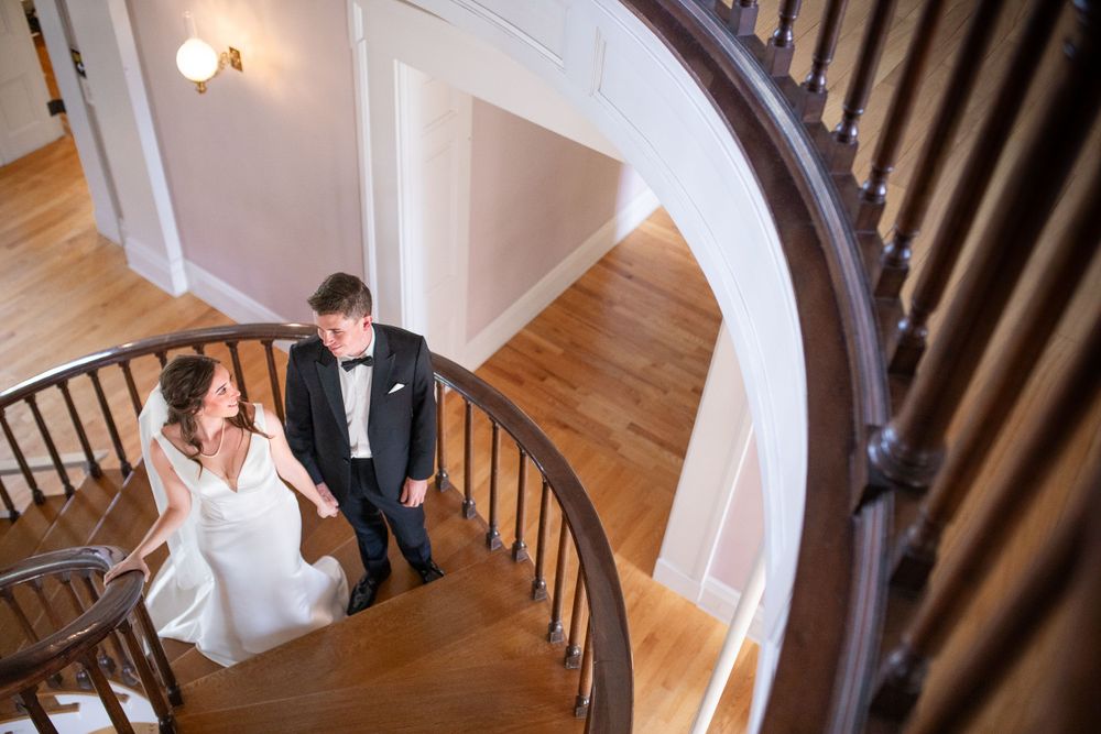 A couple in formalwear descend a curved staircase with dark wood railings in an elegant hallway.  Iowa City wedding photographer.