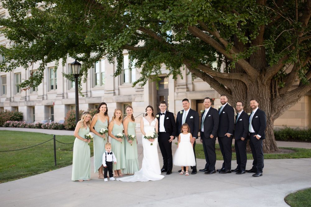 Wedding party poses under a large tree outside a light-colored stone building in elegant formal attire.  Iowa City wedding photographer.