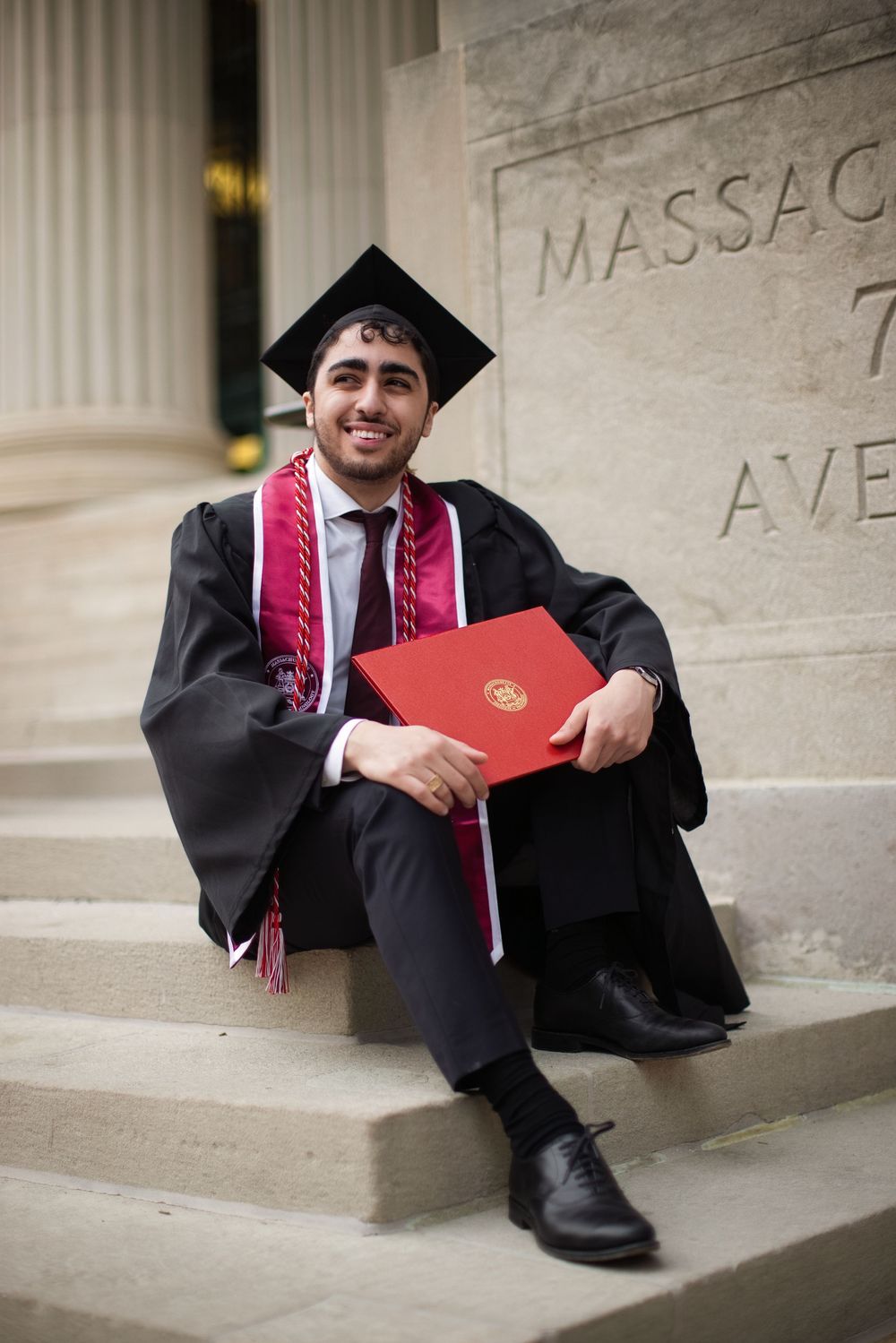 Smiling graduate in black cap and gown holds red diploma on stone steps near Massachusetts Avenue sign.