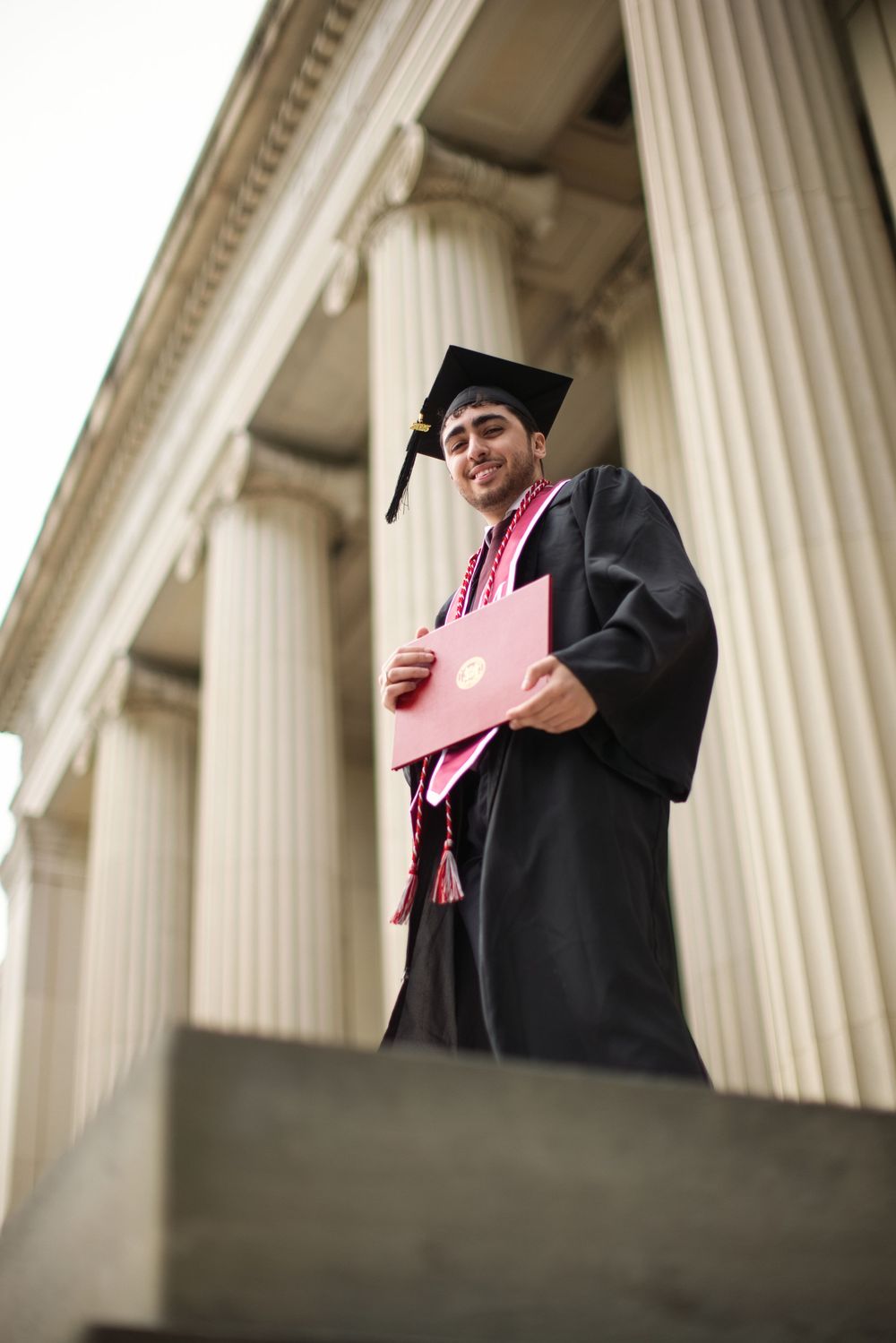 Smiling male graduate in black cap and gown holds pink diploma outside columned university building.