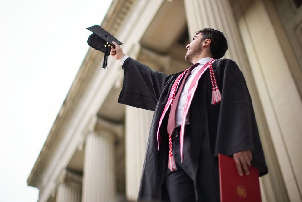 Graduate in black gown and pink sash tosses cap in the air outside a classical university building.