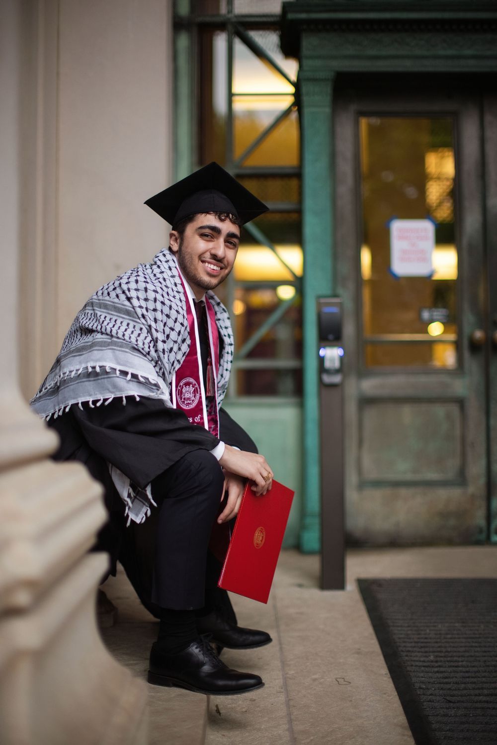 Smiling graduate in cap, gown, and keffiyeh holds red diploma folder while posing on steps outside a building.