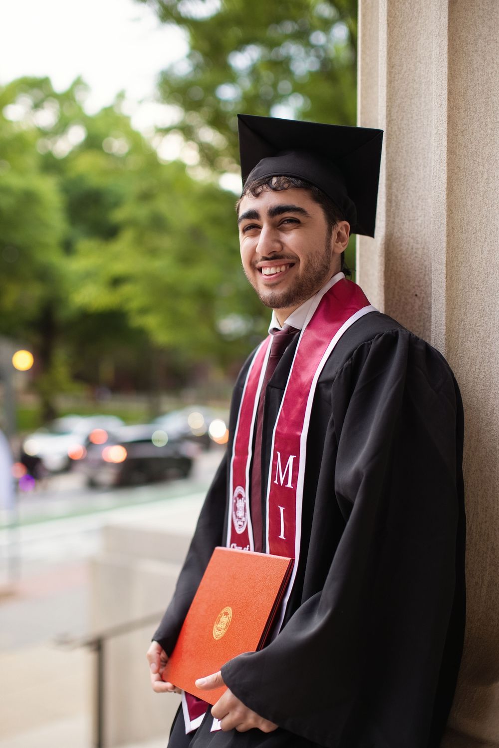 Smiling graduate in black cap and gown holding orange diploma folder, wearing red stole, leaning against column outdoors.