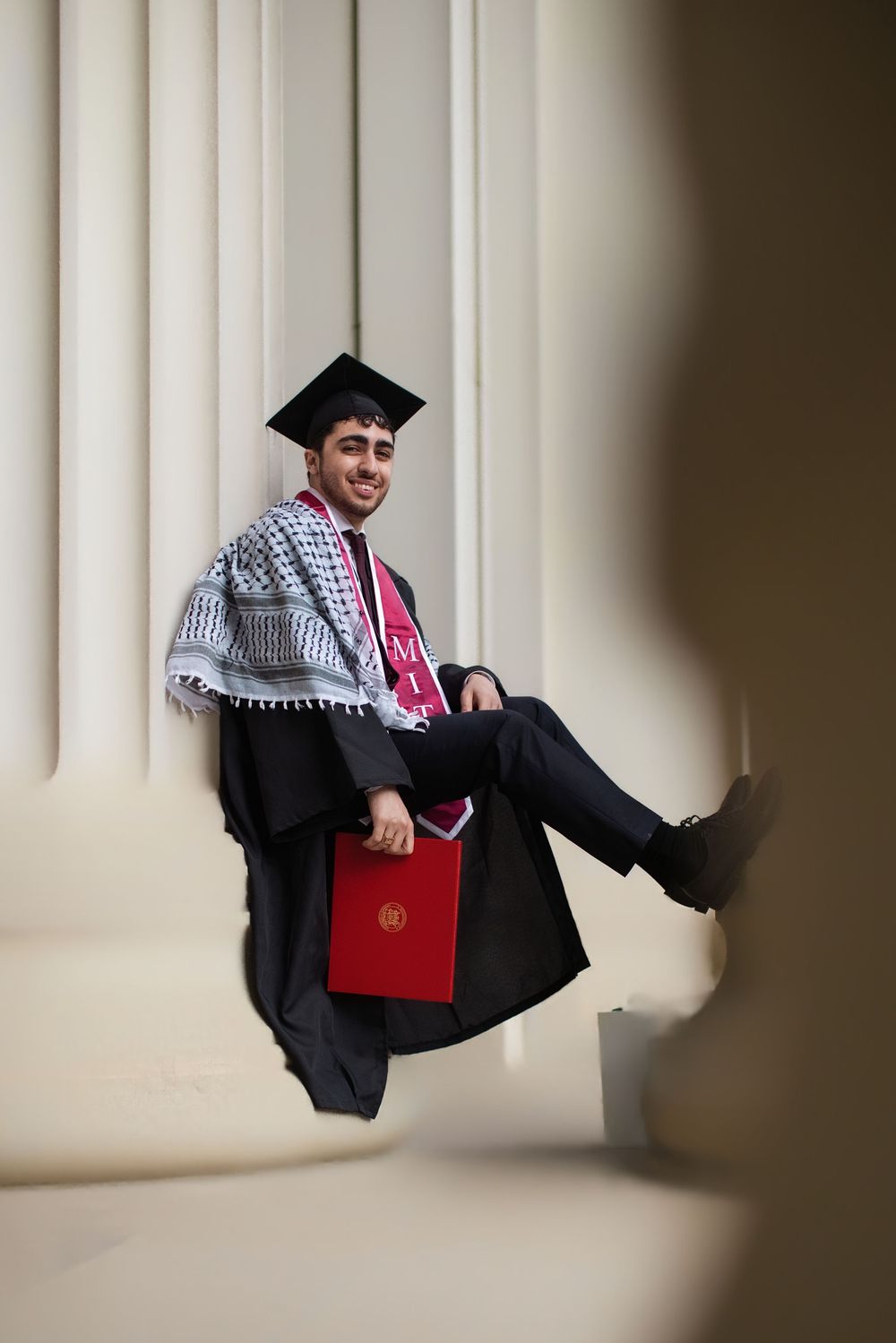 Smiling graduate in cap and gown with keffiyeh scarf holds red diploma folder while sitting between white columns.