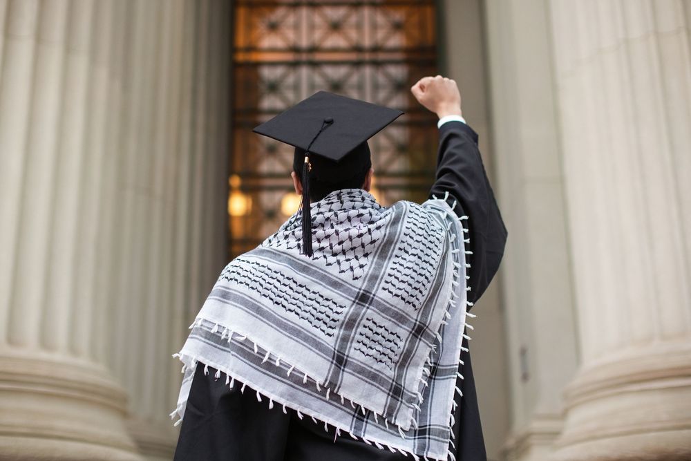 Graduate in cap and gown wearing a keffiyeh raises fist in front of a columned building.