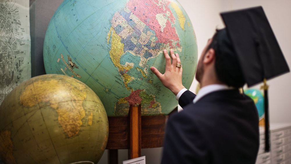 Graduate in cap and gown pointing at a large vintage globe in an academic setting.