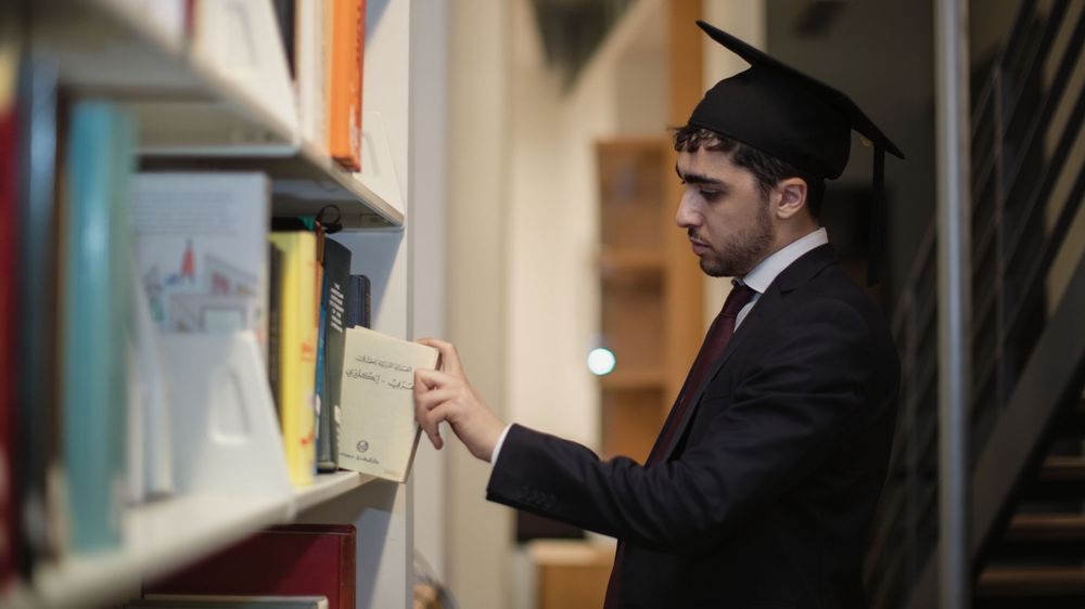 A graduate in cap and gown browses books on a library shelf, reaching for a volume among rows of colorful titles.