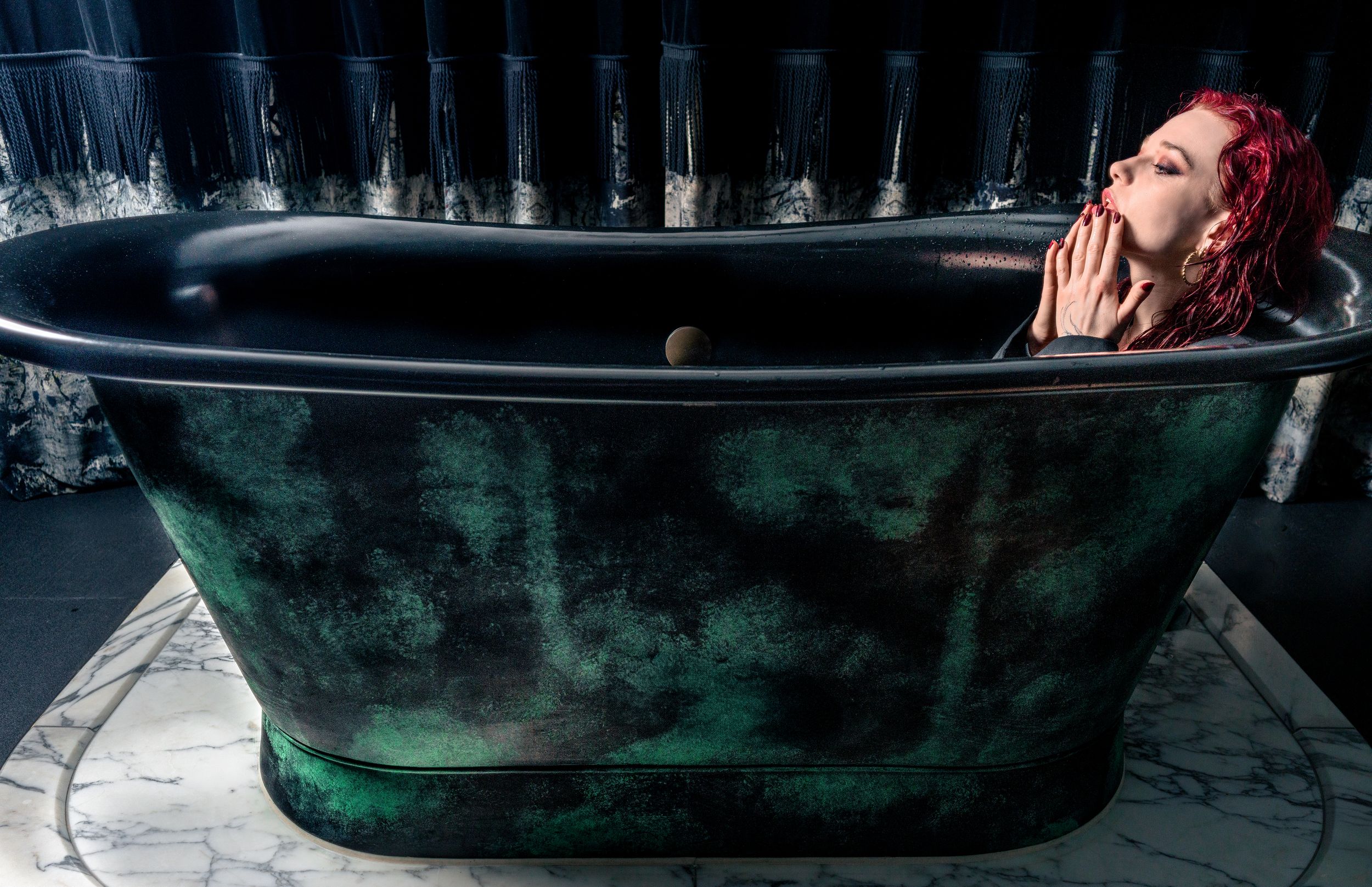 Editorial portrait of a model with red hair reclining in a dark vintage bathtub