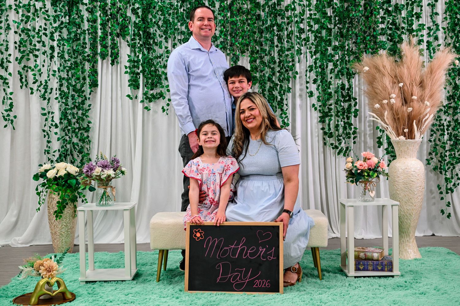 Family of four posing for a Mother's Day photo in front of a green ivy backdrop with floral decor.
