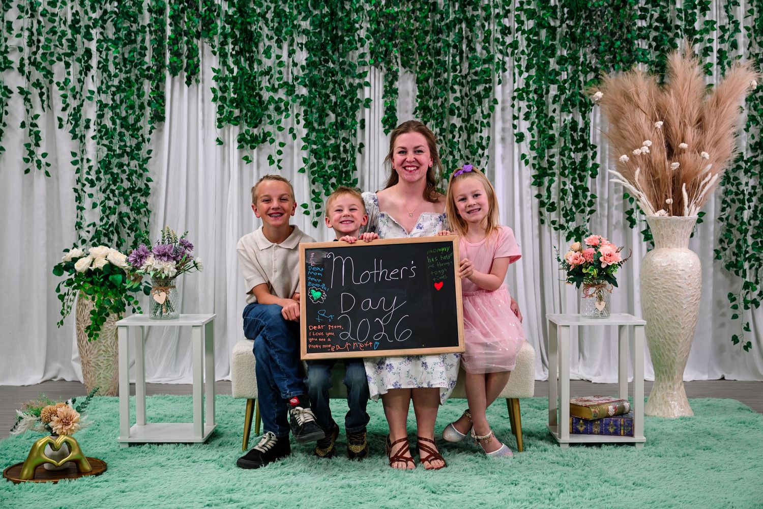 A mother poses with three children holding a Mother's Day chalkboard sign in a decorated photo booth setting.