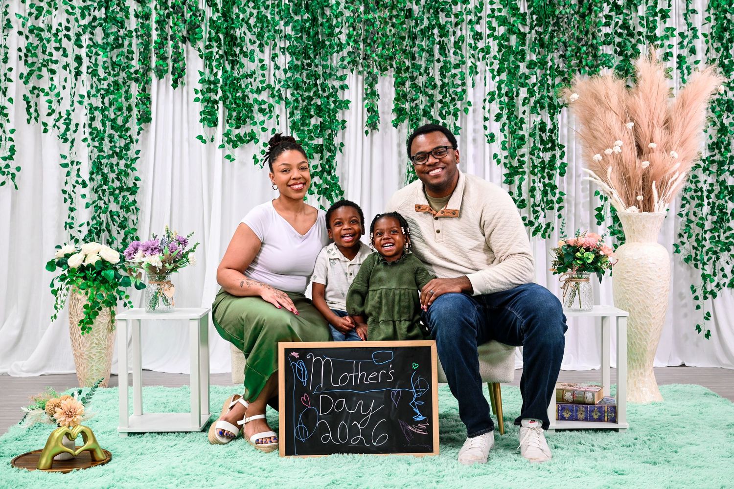 Family of four poses for a Mother's Day 2026 photo with green floral backdrop and chalkboard sign.