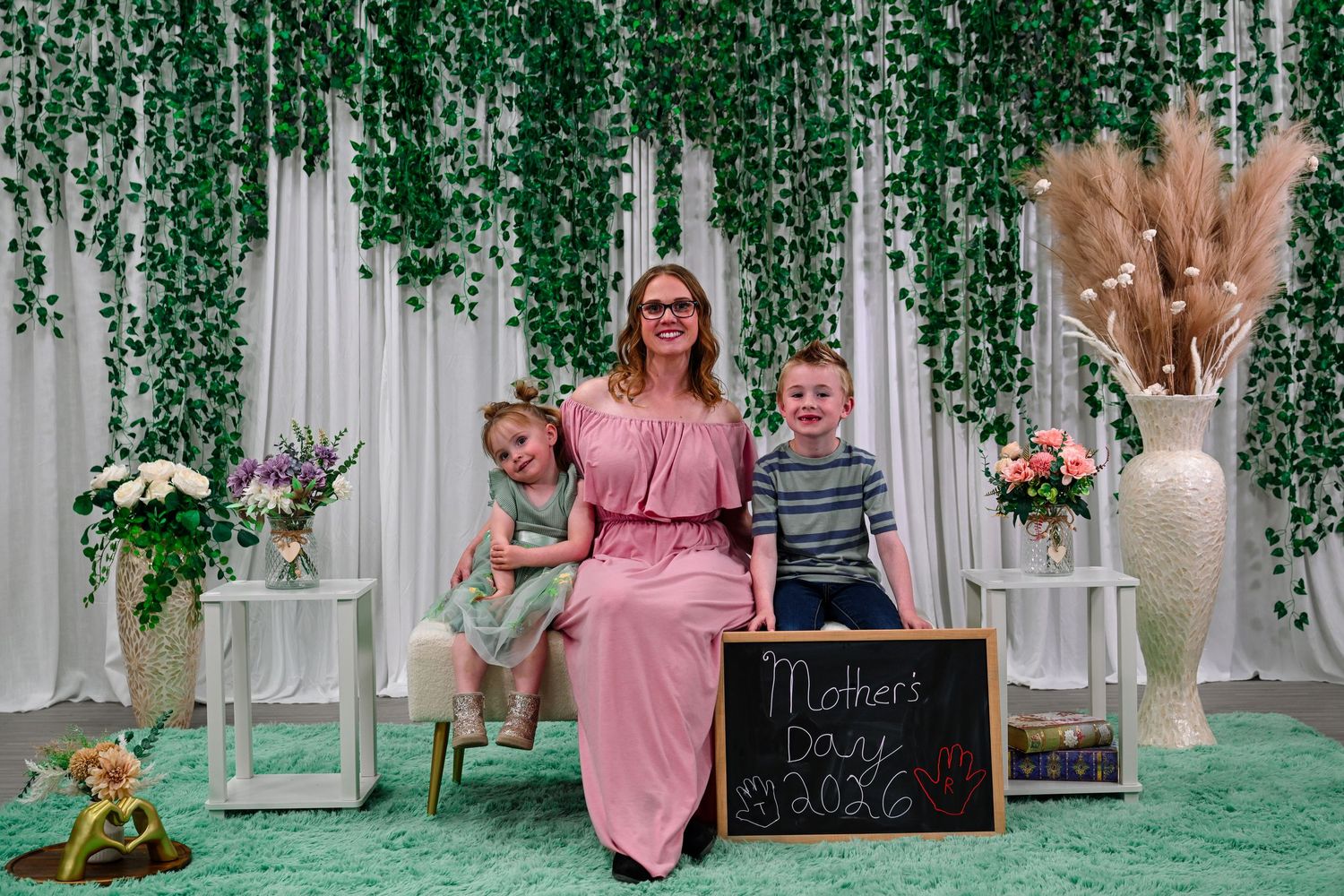A mother in a pink dress poses with two young children at a Mother's Day photo session with green ivy backdrop.
