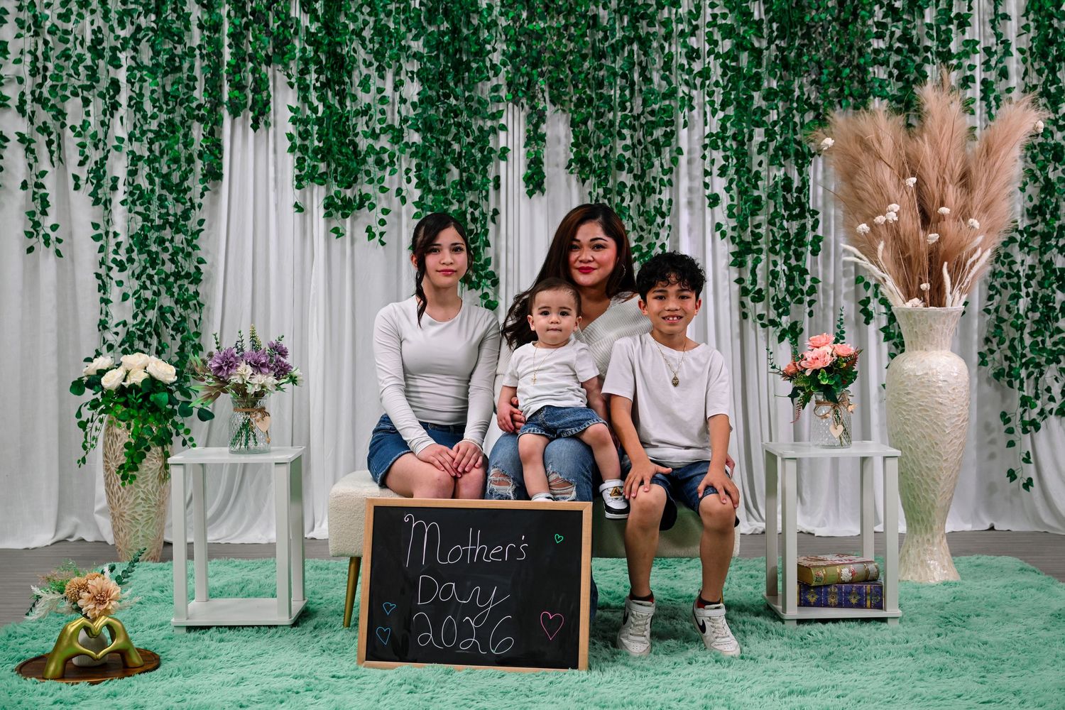 Family of four poses for a Mother's Day 2026 photo in front of a green vine backdrop with white curtains.