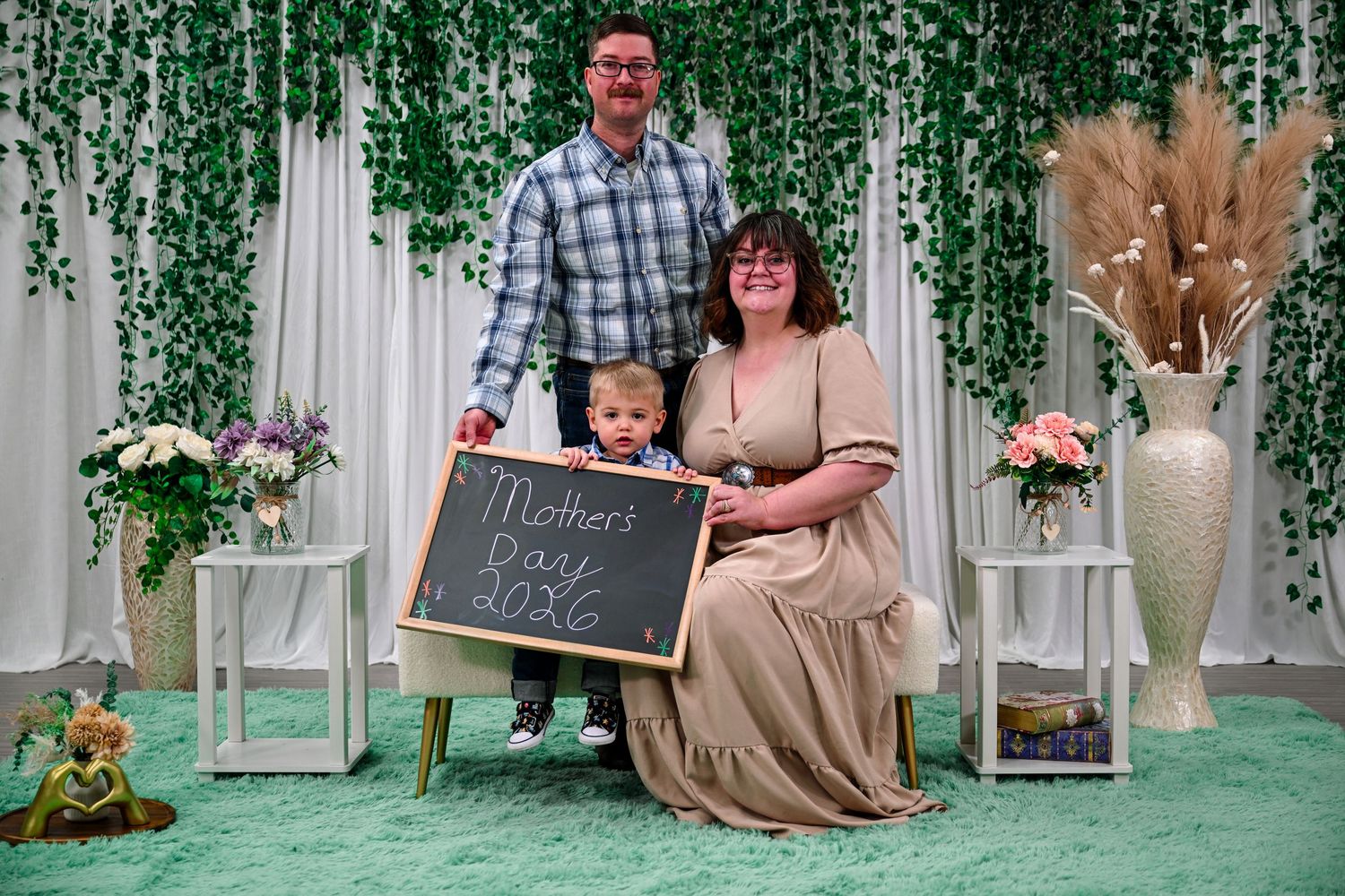 Family of three posing with a Mother's Day 2026 chalkboard sign in a decorated floral photo booth setting.