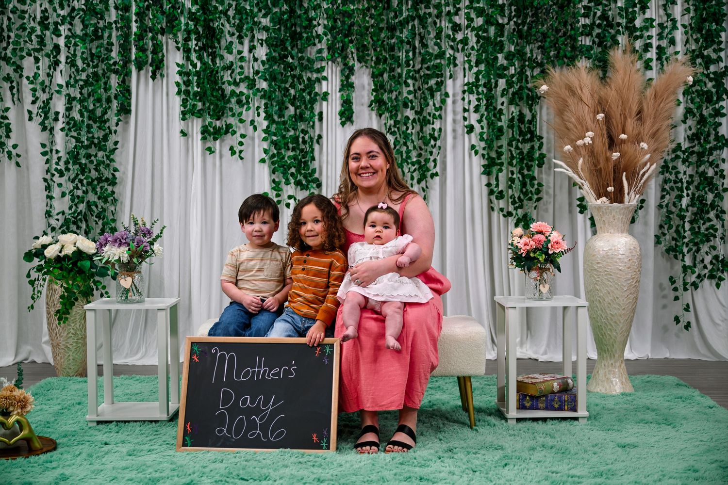 A mother poses with three young children on Mother's Day 2026 in front of a lush green ivy backdrop.