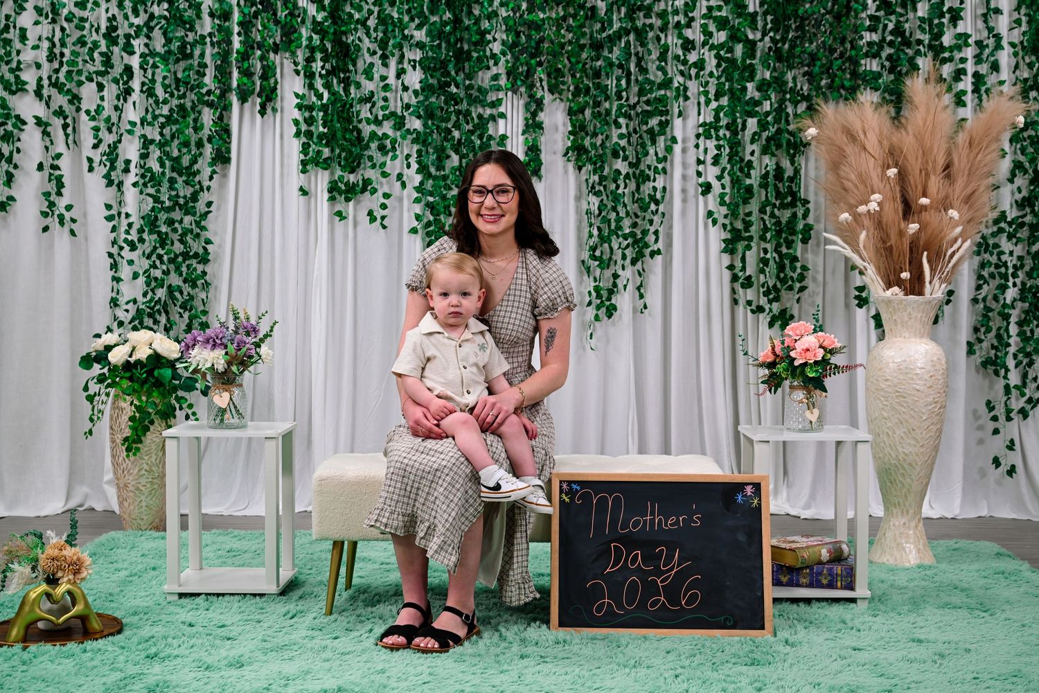Mother and toddler posing together at a Mother's Day 2026 photo backdrop with greenery and flowers.