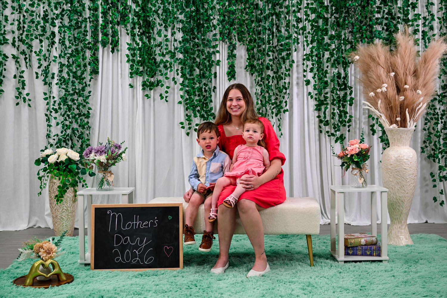 A mother in a red dress poses with two young children on a bench at a Mother's Day 2026 photo backdrop.