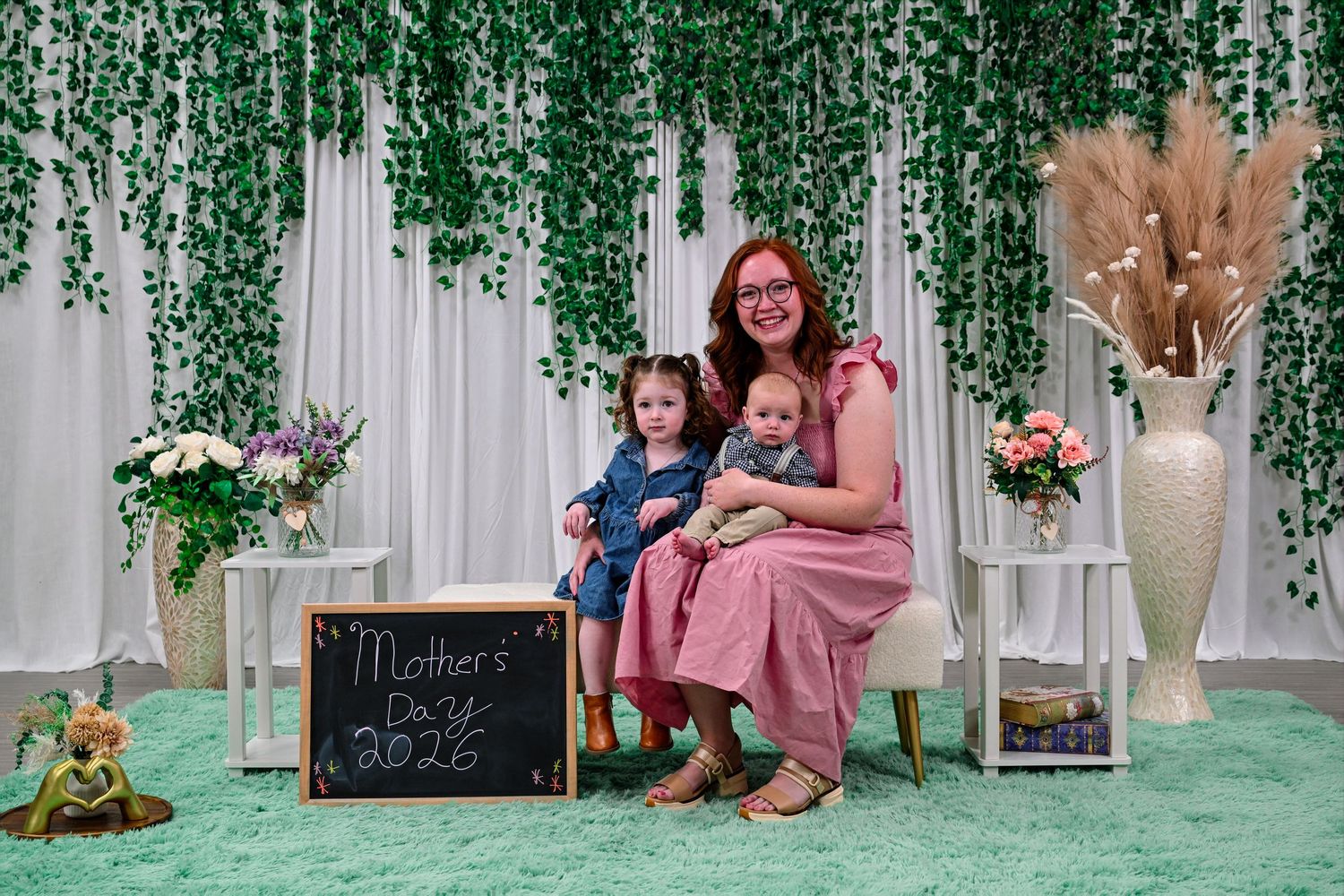 Mother in pink dress poses with two young children for a Mother's Day 2025 photo with floral backdrop.