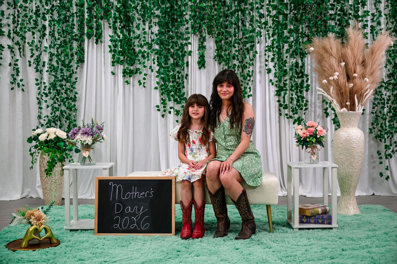 A woman and young girl pose together in front of a green ivy backdrop with a Mothers Day 2026 chalkboard sign.