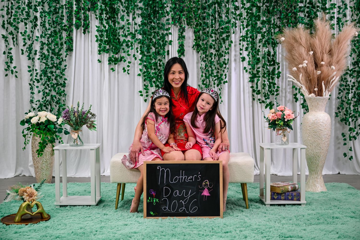 A mother poses with two daughters in pink outfits holding a Mother's Day 2026 chalkboard sign against a green backdrop.