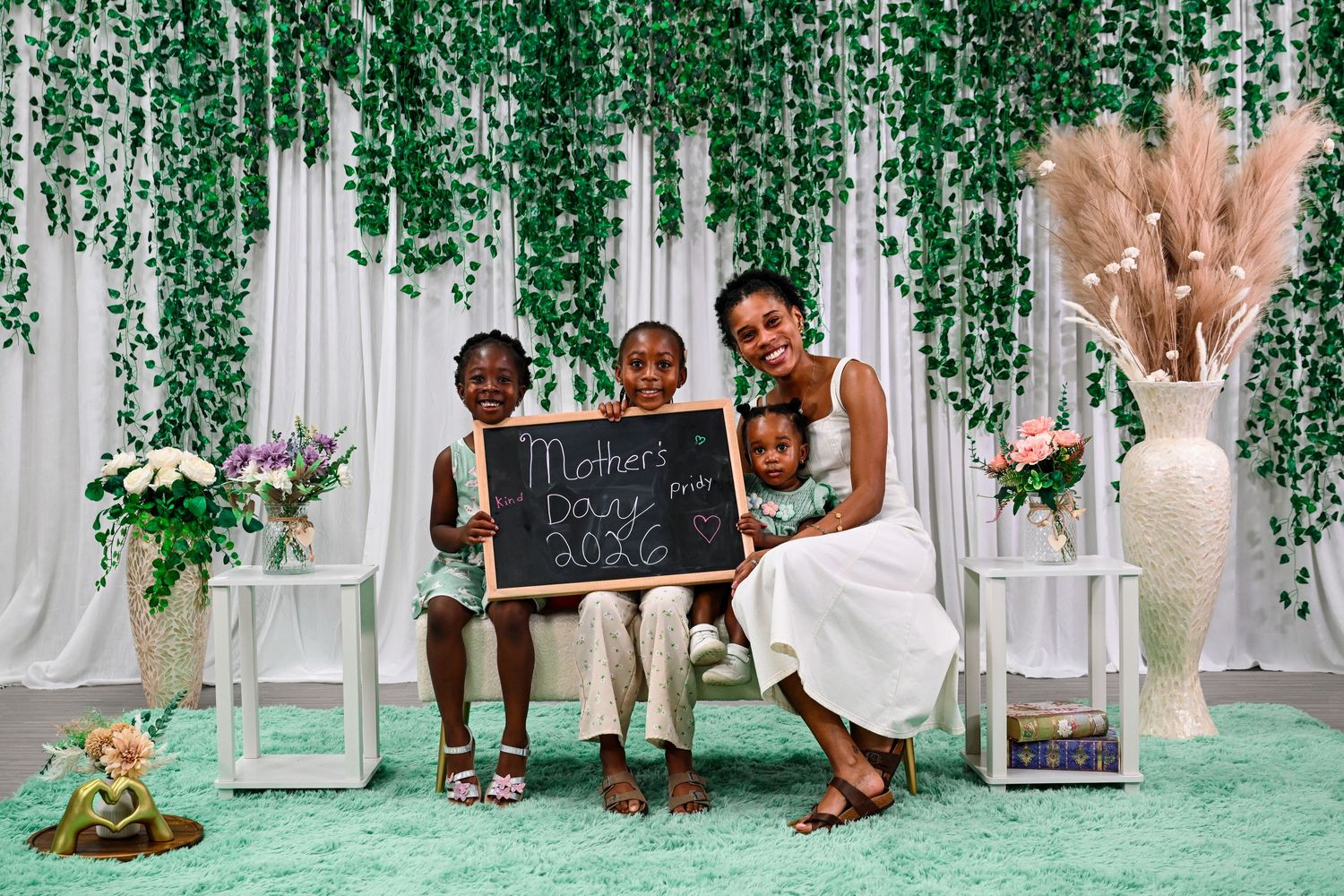 A mother and three children pose with a Mother's Day 2023 chalkboard sign amid green floral decor.