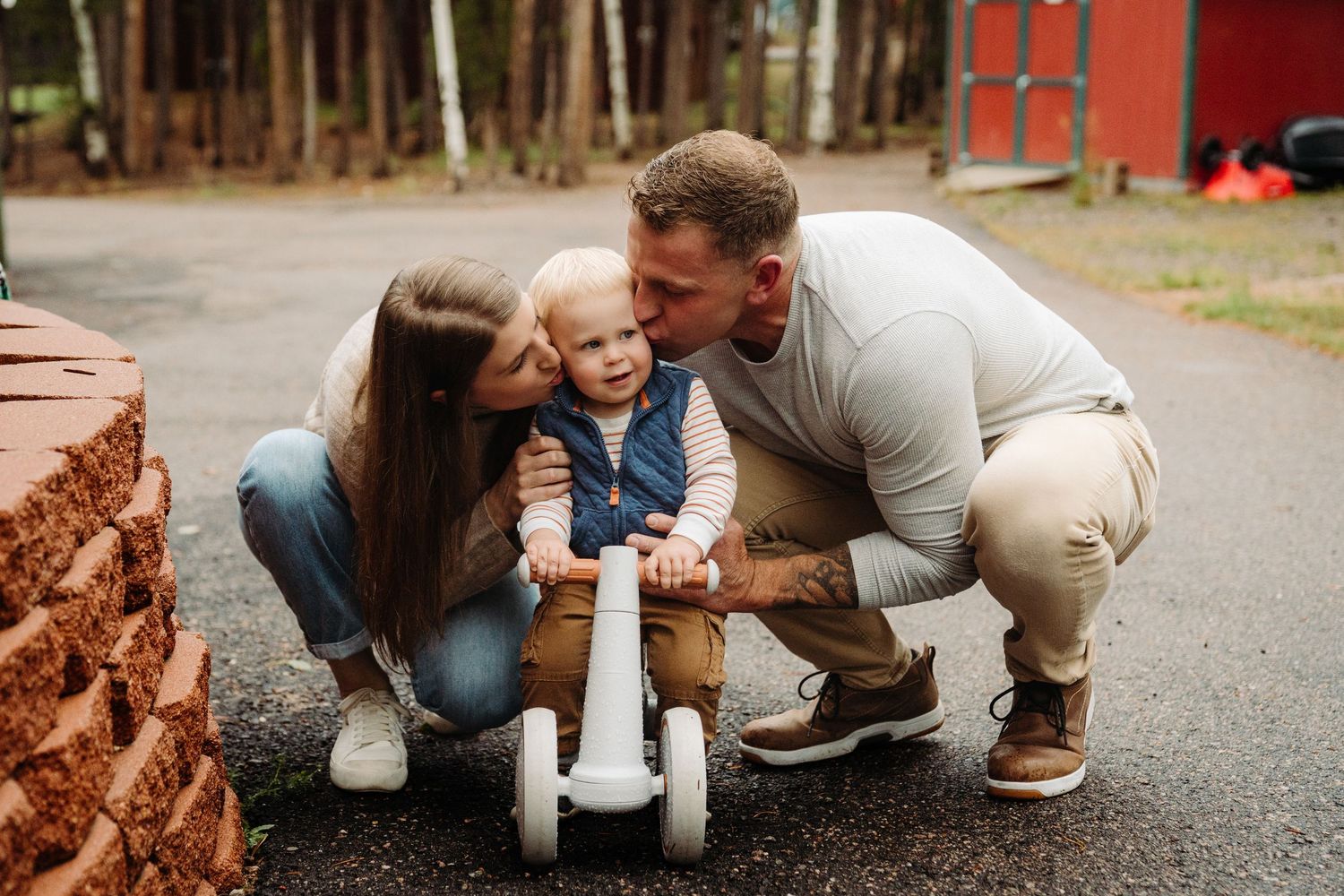 Parents kissing their baby boy sitting on a toy tricycle outdoors near brick wall and pine trees.