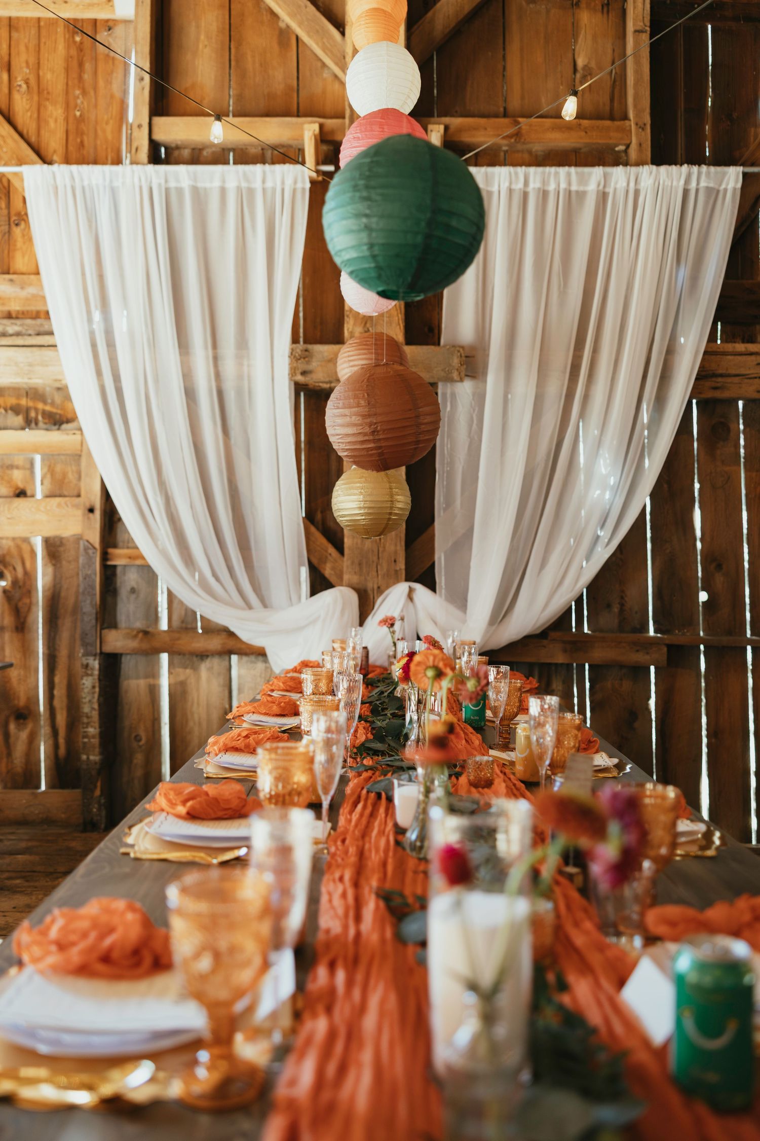 Rustic barn wedding reception table with orange linens, candles, and colorful paper lanterns hanging from white draped fabric.