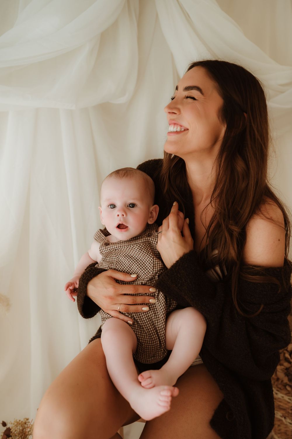 Smiling mother holding her baby in a brown onesie against soft white draped fabric backdrop.