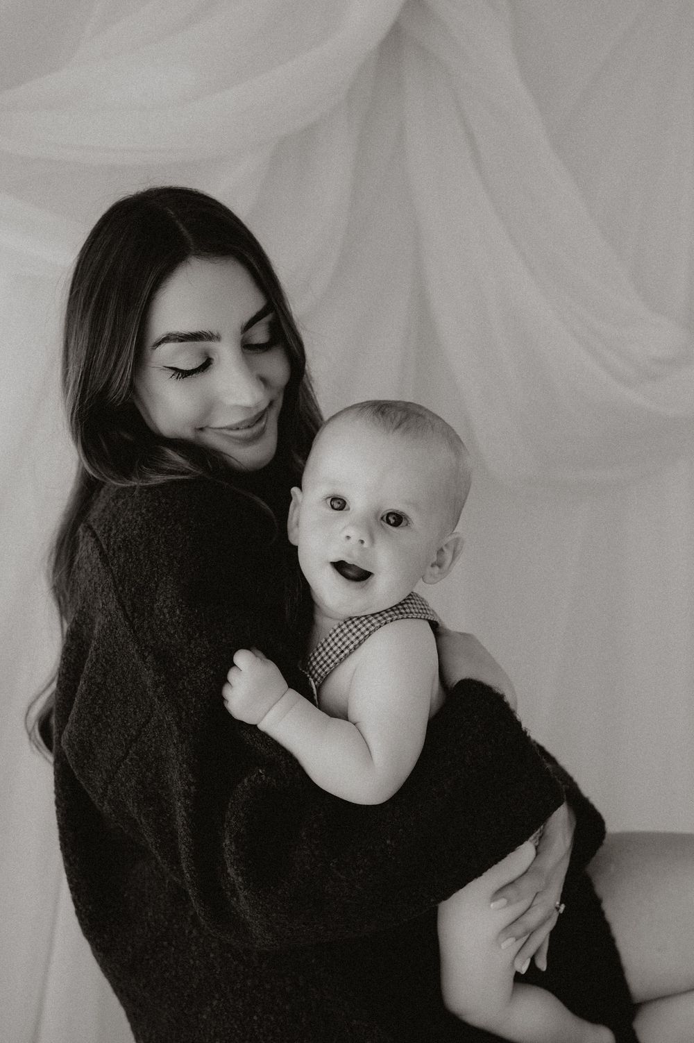 Black and white photo of a smiling mother holding her happy baby in a checkered romper against soft draped fabric.