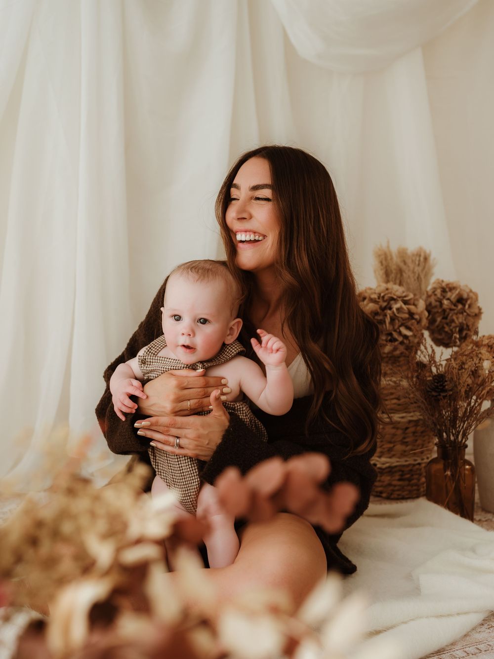 A smiling woman with long dark hair holds a baby in a boho-styled studio with dried florals and neutral tones.