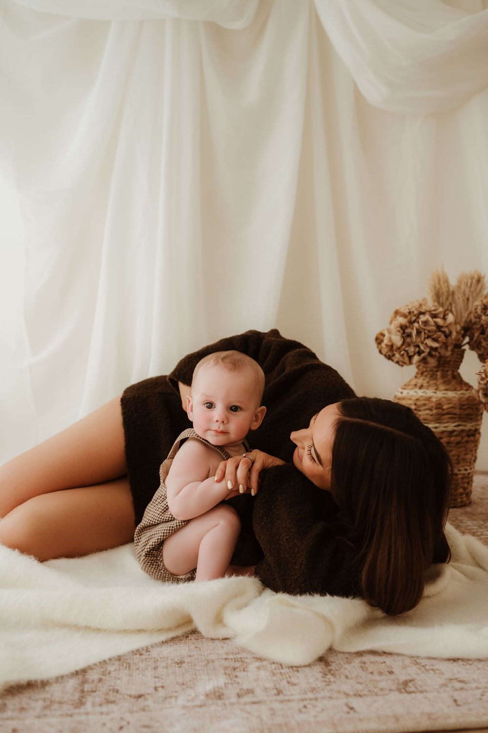 A mother in a dark outfit lies on a white blanket gazing at her baby in a boho studio setting.
