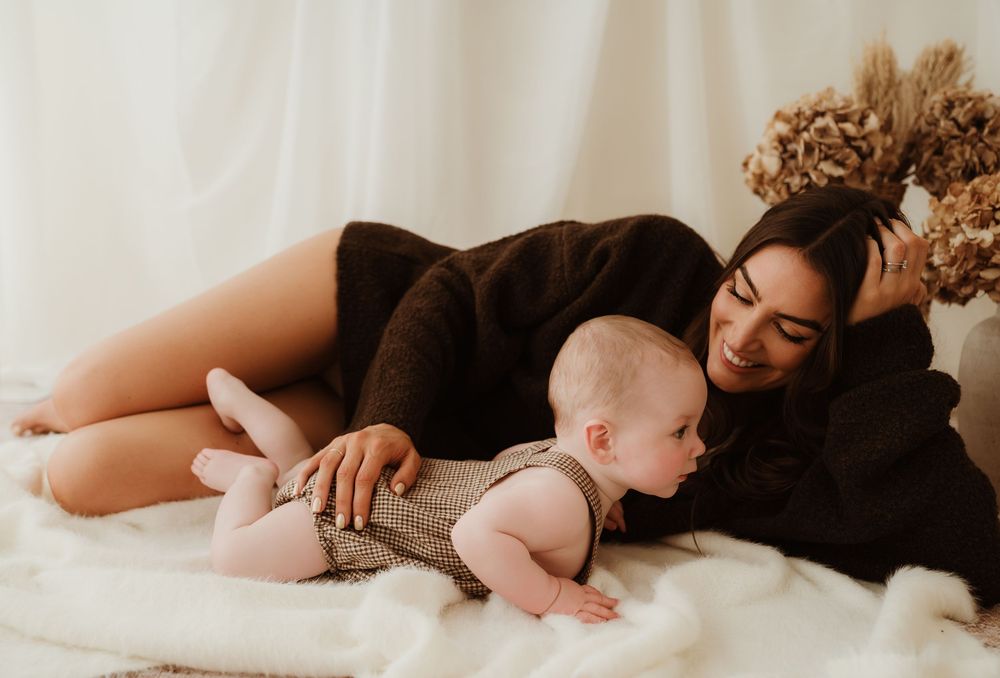 A mother in a brown outfit lies on a white blanket, smiling at her crawling baby in a cozy indoor setting.