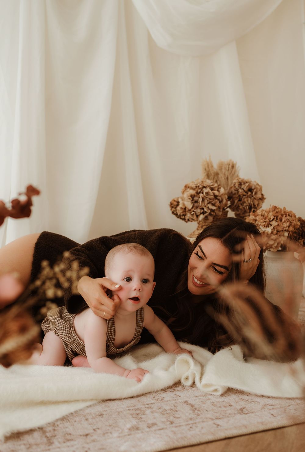 A smiling mother lies beside her curious baby on a white blanket with dried floral decor behind them.