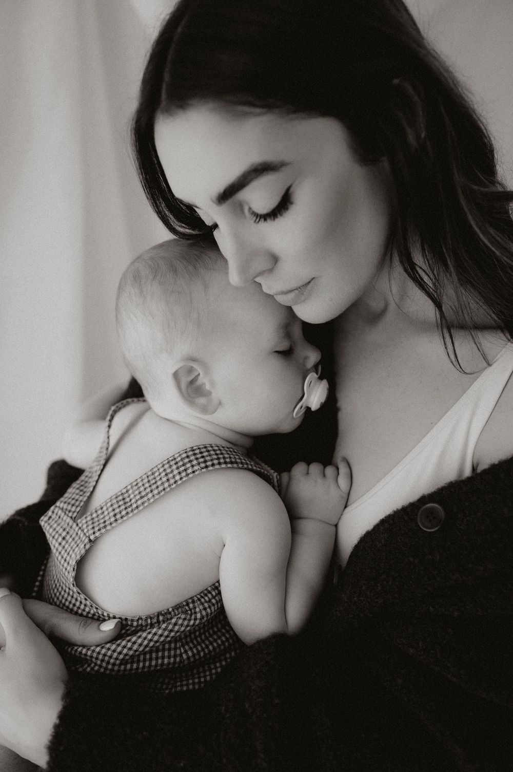 Tender black and white portrait of a dark-haired mother gently kissing her baby in a checkered romper.