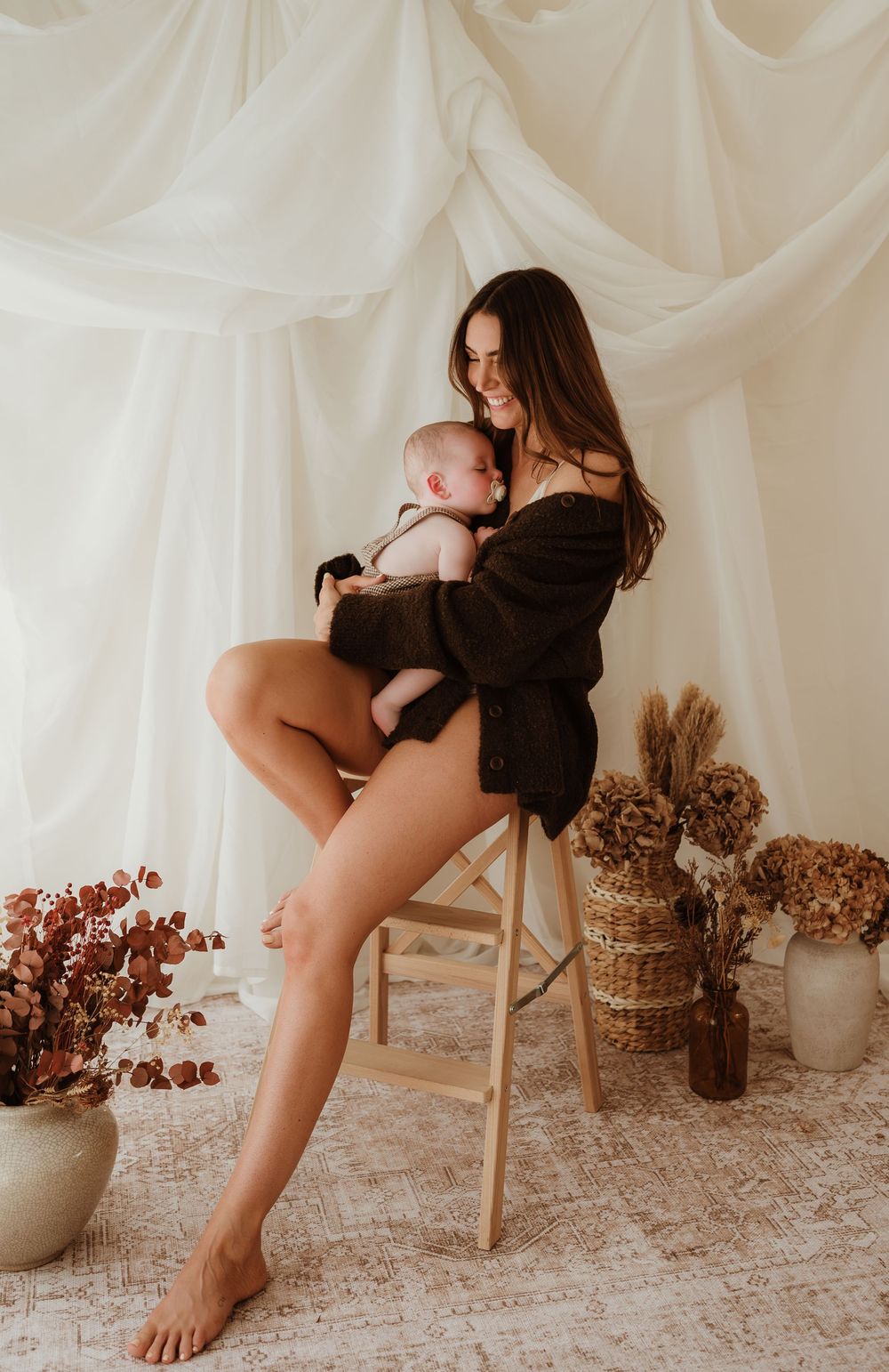 Mother sitting on wooden stool cradling newborn baby in dark robe surrounded by dried floral arrangements.