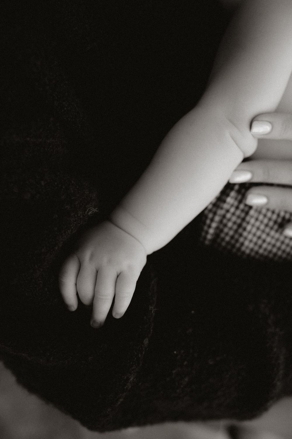 Black and white photo of a tiny baby hand resting gently, with an adult's manicured fingers visible nearby.