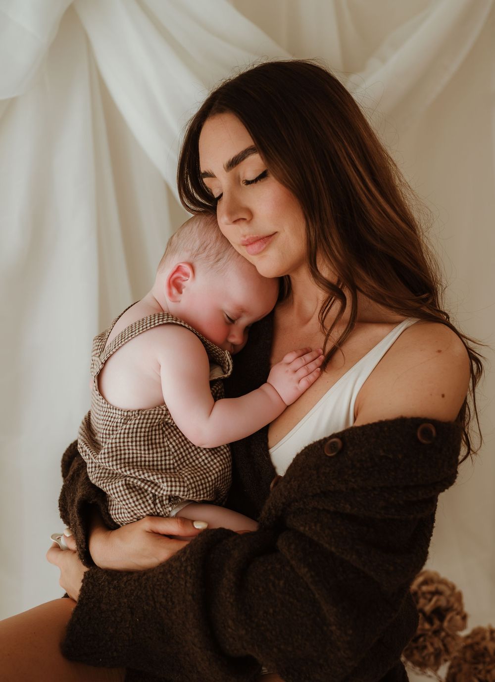 A young mother with long dark hair tenderly holds her sleeping newborn baby, both sharing a peaceful moment.