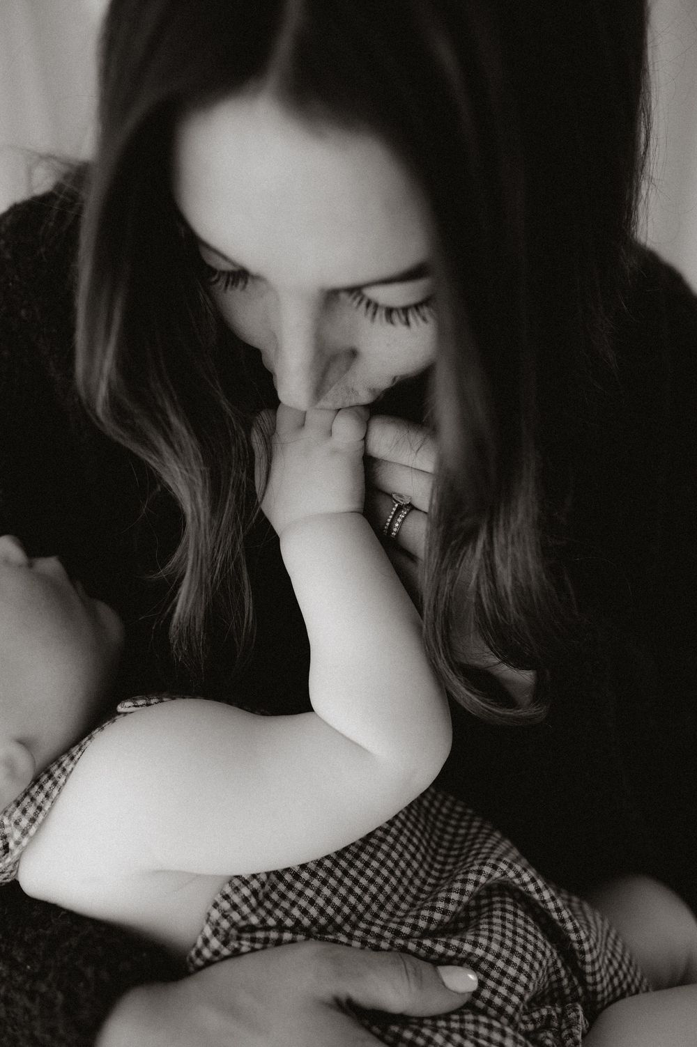 Tender black and white portrait of mother kissing baby hand while holding infant close.