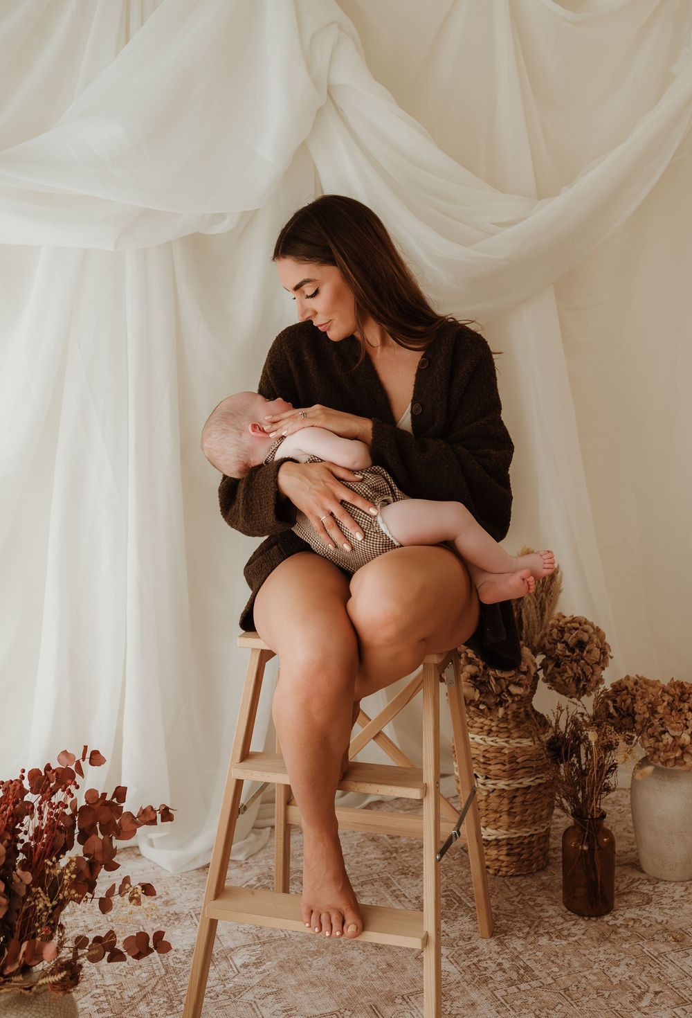 Loving mother gazing down at sleeping baby on wooden ladder stool with dried flowers in background.