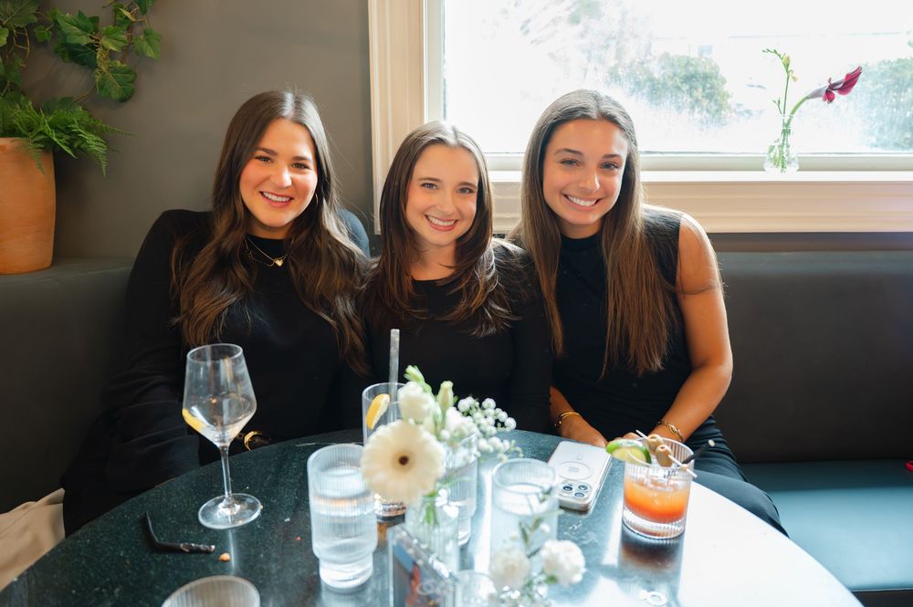 Three young women in black outfits smile together at a restaurant table with drinks and flowers.