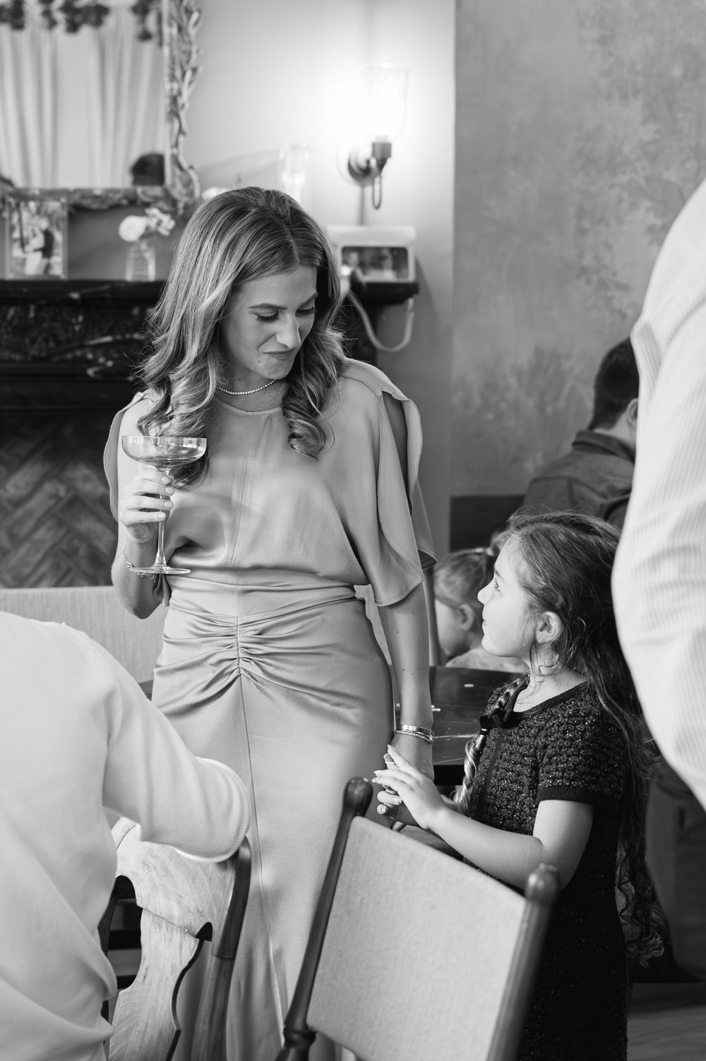 Black and white photo of an elegant woman holding wine glass conversing with a young girl at a formal event.