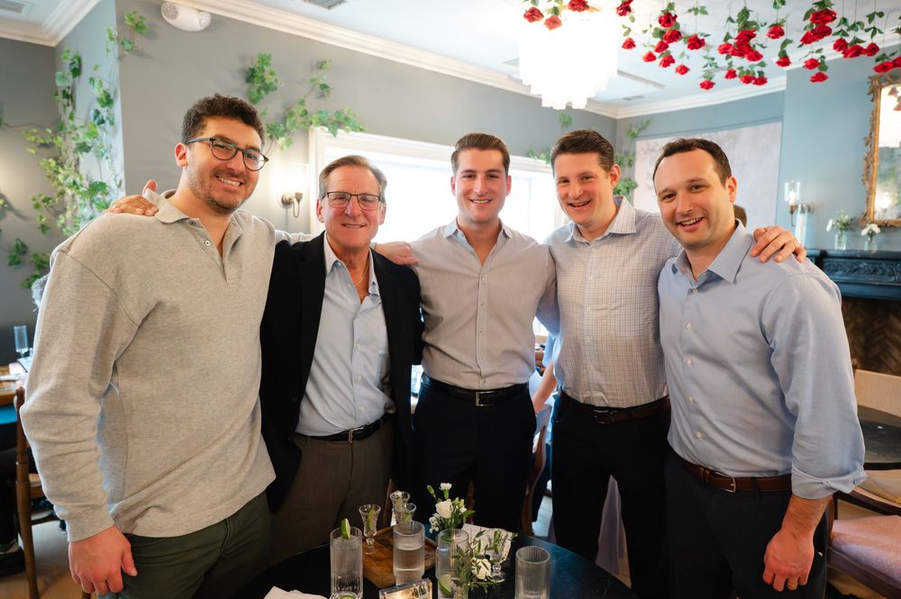 Five smiling men pose together in an elegant restaurant with floral decor and soft lighting.